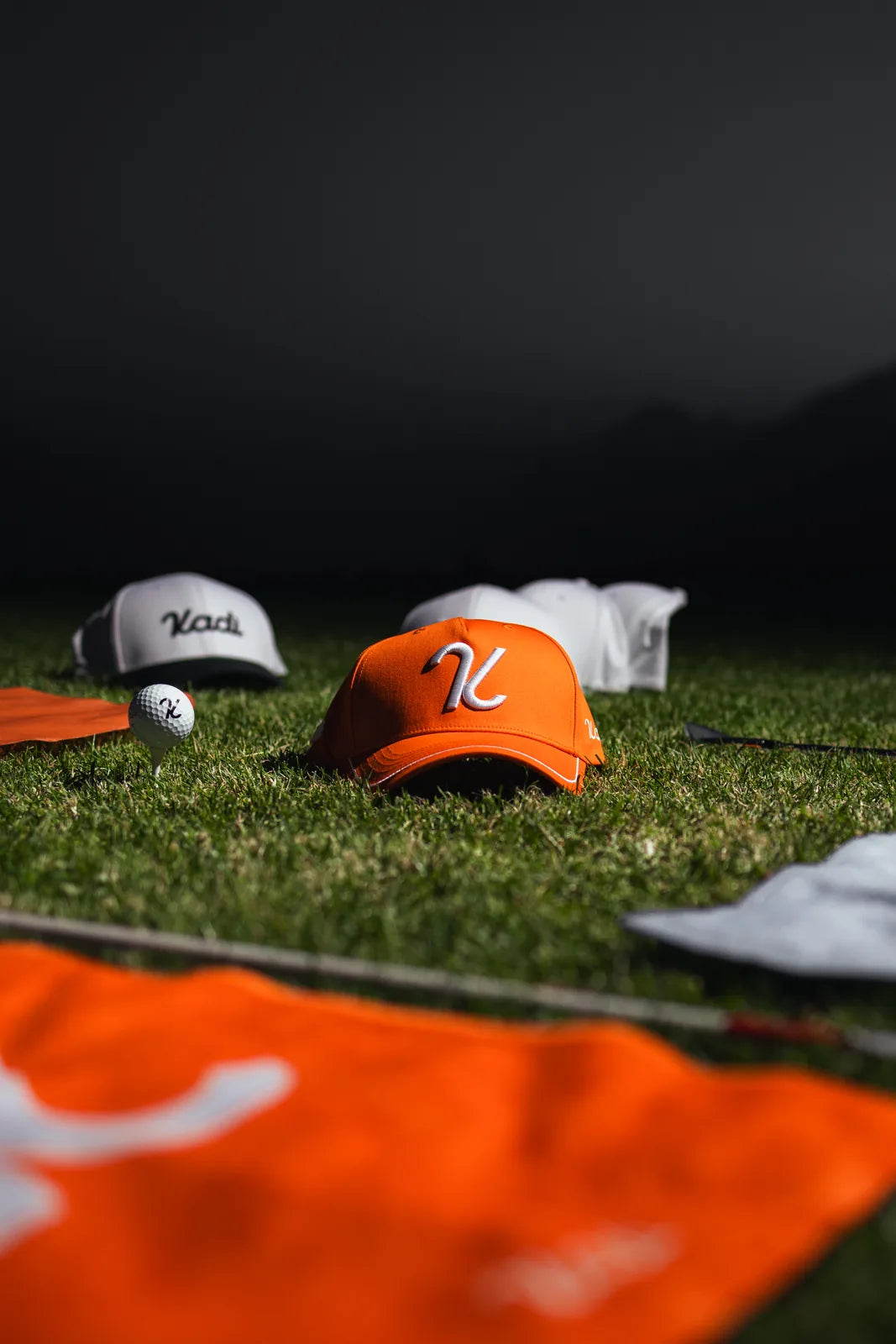 KADI caps in white and orange with a golf ball on the grass at dusk