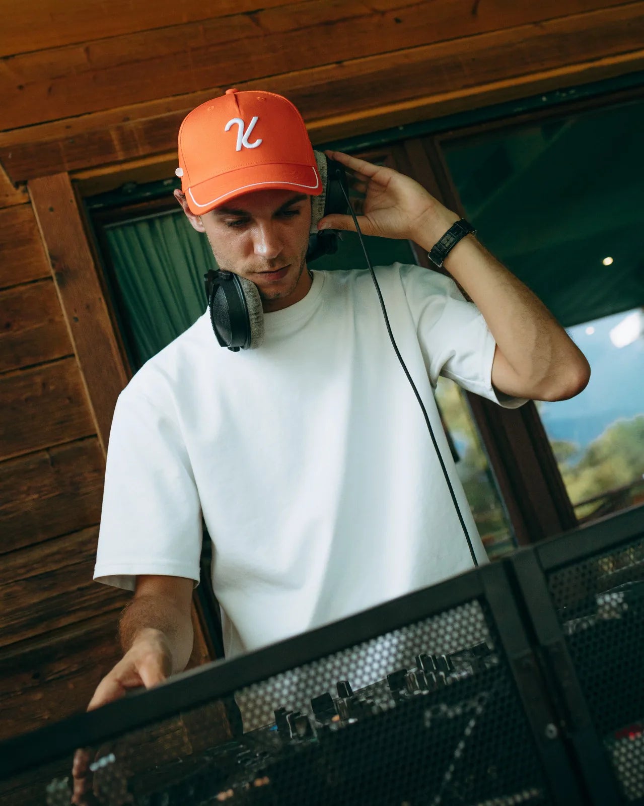 Man in an orange KADI cap and studio headphones at a DJ mixer by a wooden chalet